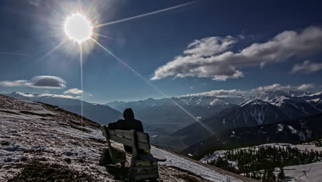 Time-lapse-shot-of-person-sitting-on-bench-of-snowy-mountaintop-and-enjoying-mountain-panorama-view-during-sunny-day-on-blue-sky-in-winter
