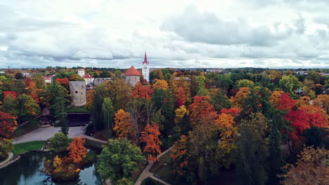 Aerial-view-of-Cēsis-Castle,-one-of-the-most-iconic-and-best-preserved-medieval-castle-in-Latvia-with-autumnal-forest-surrounding-the-fortress-on-a-cloudy-day