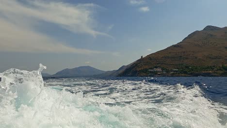 Low-angle-water-surface-slow-motion-pov-of-boat-wake-trail-with-Riserva-dello-Zingaro-natural-reserve-of-Sicily-in-background