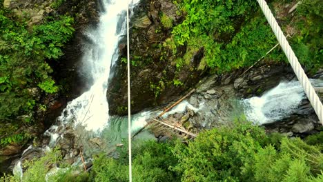 Eine-POV-Aufnahme-Eines-Weißen-Mannes-In-Blauen-Stiefeln,-Der-über-Eine-Stahlseilbrücke,-über-Einen-Wilden-Fluss-Und-Einen-Wasserfall-Balanciert
