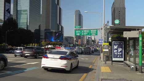 Police-Car-Passes-by-empty-Bus-Stop-during-heavy-traffic-at-Samseong-Station,-COEX,-Seoul-High-rise-Skyscraper-Building-on-the-background-when-cards-drive-on-Yeongdong-road