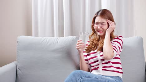 Asian-young-woman-sitting-on-the-sofa-drinking-water-to-relax-during-severe-headache