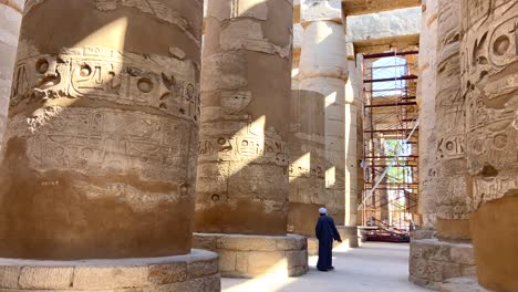 Local-Egyptian-person-with-tunic-and-turban-greeting-and-walking-between-huge-columns-of-Karnak-temple-under-construction-in-Luxor-City,-Egypt