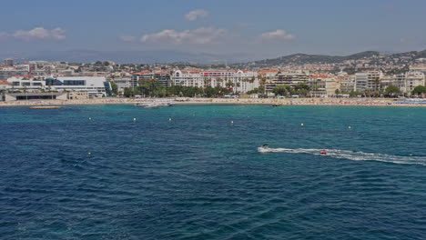 Cannes-France-Aerial-v39-establishing-low-level-pan-shot-overlooking-croisette-beach-and-cityscape,-with-speed-boats-sailing-and-water-activities-in-the-foreground---July-2021