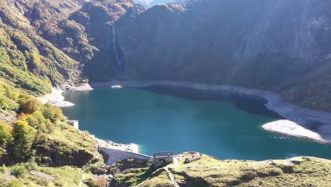 Lac-d'Oô-artificial-lake-in-the-French-Pyrenees-with-waterfall-and-dam-wall,-Aerial-lowering-reveal-shot