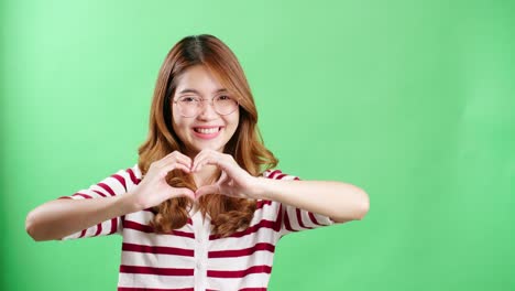 Junge-Asiatische-Frau-Mit-Brille,-Die-Im-Studio-Mit-Chromakey-Ein-Herzsymbol-Mit-Den-Händen-Macht