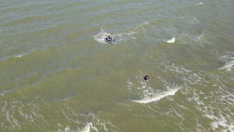 A-descending-aerial-footage-of-a-man-wading-towards-the-shore,-a-Jet-Ski-waiting,-waves-rushing