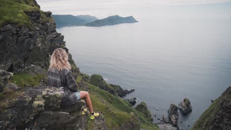 Blonde-woman-with-yellow-sneakers-sitting-on-lands-end-in-Runde-island,-Norway