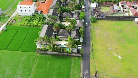 cars-and-motorbikes-driving-on-road-through-Berawa-Bali-next-to-green-rice-fields-and-villas,-aerial