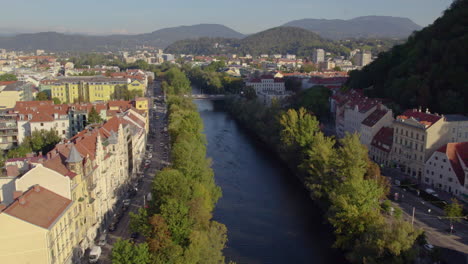Luftaufnahme-Entlang-Des-Malerischen-Flusses-Mur,-Leuchtend-Rote-Dachgebäude-Am-Ufer-Des-Schlossbergs,-Österreich