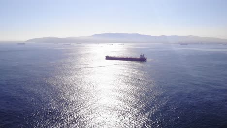 Luftaufnahme-Eines-Frachtschiffs,-Das-Durch-Die-Straße-Von-Gibraltar-Fährt,-Mit-Sonnenschein,-Der-Sich-Auf-Der-Wasseroberfläche-Spiegelt