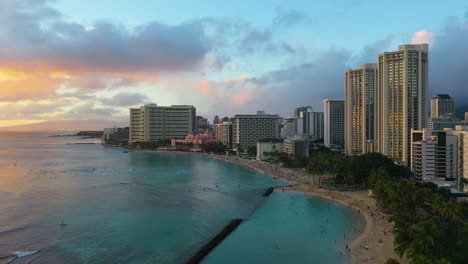 Vista-Aérea-De-Drones-De-Los-Edificios-Del-Hotel-Resort-Frente-Al-Mar-Y-Gente-Nadando-En-La-Playa-De-Kuhio-En-Waikiki,-Hawaii-Al-Atardecer-Con-Muro-De-Ruptura