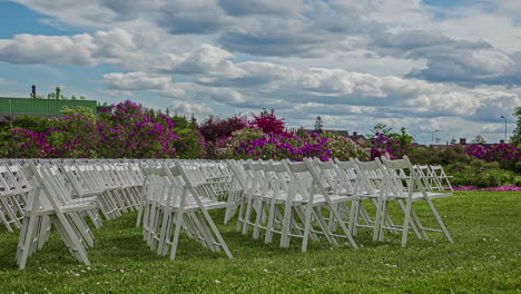 Static-shot-of-rows-of-wooden-folding-white-chairs-on-the-green-grass-garden-floor-with-white-clouds-passing-by-on-a-cloudy-day-in-timeplase