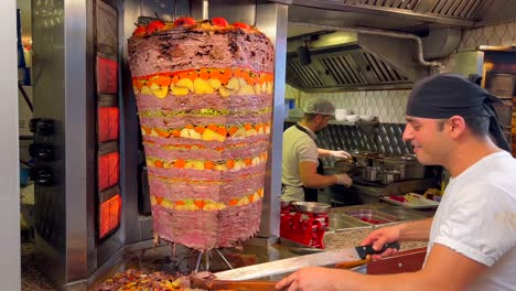 Happy-smiling-man-cutting-meat-from-a-vertical-rotisserie-at-a-kebab-restaurant-in-Bodrum-Turkey,-people-working-in-the-kitchen,-good-traditional-food,-seasoned-meat-with-potatoes-and-carrots,-4K-shot