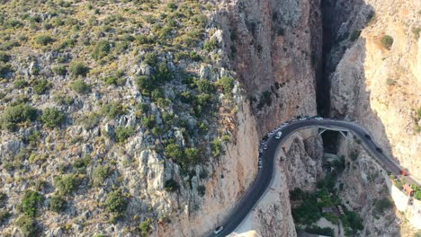 aerial-drone-panning-upwards-as-cars-travel-on-a-coastal-road-alongside-a-steep-mountain-cliff-at-Kaputas-Beach-in-Kas-Turkey-revealing-the-large-mountain-above-on-a-sunny-summer-day