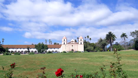 Reveal-shot-of-the-picturesque-Santa-Barbara-Mission-building-from-behind-bright-red-roses-in-California