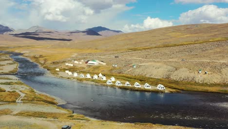 Aerial-drone-panning-across-a-natural-river-with-white-luxury-glamping-tents-in-the-high-altitude-alpine-plain-of-Deosai-National-Park-located-between-Skardu-and-Astore-Valley-in-Pakistan-in-summer