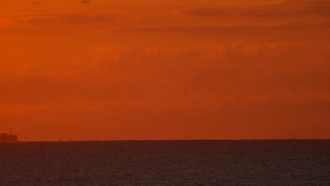 A-cargo-ship-travels-across-in-the-distance-across-the-ocean-horizon-under-a-glowing-orange-sky