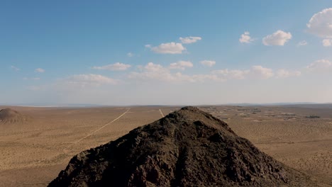 Rocky-mountaintop-in-vast-Mojave-Desert-plain-landscape,-Aerial-Pull
