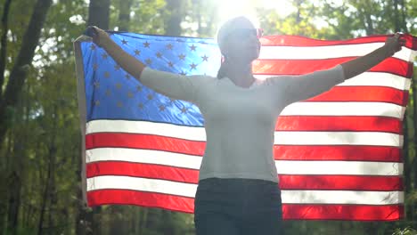 Closeup-of-backlit,-pretty-blonde-woman-walking-through-a-forest-holding-a-flag-up-in-the-air-behind-her