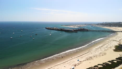 Aerial-shot-flying-out-above-a-sandy-California-beach-with-Stearns-wharf-pier-and-the-clear-blue-ocean-harbor-full-of-sail-boats