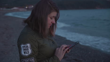 A-girl-wearing-a-jacket-uses-her-phone-on-the-beach-on-the-sunset-in-autumn