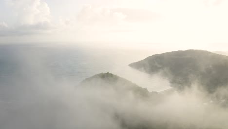 Beautiful-drone-approaching-through-the-clouds-a-peak-of-a-tropical-forest-mountain-hiking-spot-in-a-summer-day-with-a-beach-and-ocean-on-the-background-at-sunrise