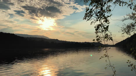 Wunderschöner-See-Mit-Silhouettierten-Bäumen-Und-Einem-Goldenen-Sonnenuntergang-In-Einem-Bewölkten-Himmel