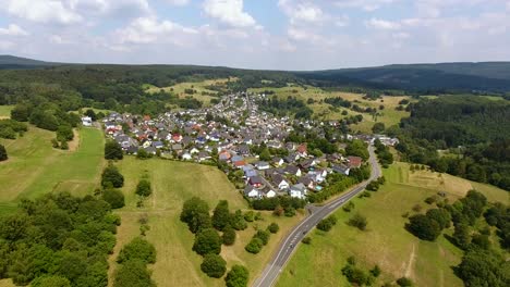 Flight-with-a-DJI-Phantom-4-Drone-towards-a-rural,-german-village-located-in-a-beautiful-forest,-hills-and-field-area