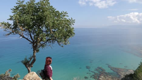 A-woman-stands-on-the-edge-of-a-mountain-beside-a-tree-looking-out-over-beautiful-blue-sea-water