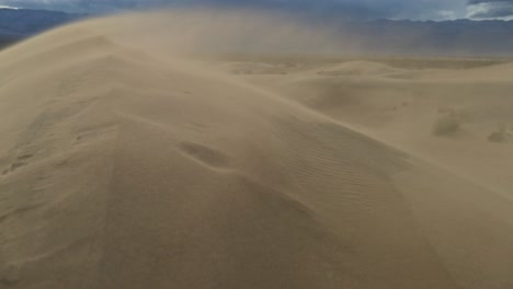 Slow-motion-sand-blowing-over-the-ridge-of-a-sand-dune,-panning-from-the-valley-to-the-peak-of-the-dune