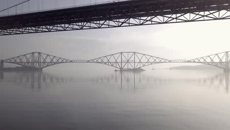 Aerial-footage-underneath-the-old-Forth-Road-Bridge-with-the-Forth-Railway-Bridge-in-the-background-on-a-sunny-day-at-South-Queensferry-in-West-Lothian,-Scotland