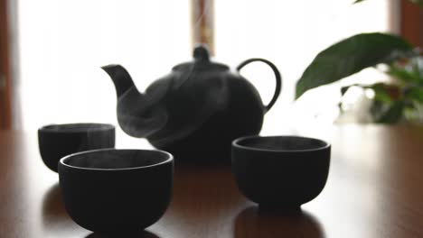 Minimaler-Hintergrund-Eines-Grünen-Japanischen-Teesets-Mit-Dampf-Aus-Den-Tassen,-Auf-Einem-Holztisch,-Mit-Einer-Pflanze-Und-Einem-Fenster-Im-Hintergrund