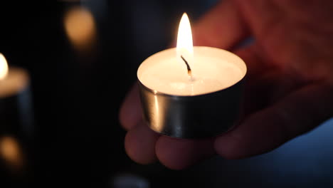 A-hand-holding-a-small-tealight-candle-with-the-flames-of-many-burning-in-the-background-during-a-cande-light-vigil-to-mourn-the-loss-of-a-loved-one