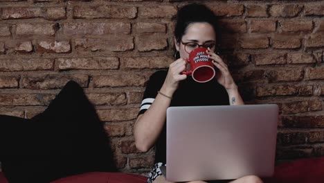 Medium-shot-of-Young-beautiful-brunette-sitting-on-bed-wearing-glasses-watching-computer-and-drinking-coffee-of-a-cup-with-the-inscription-"Just-one-more-episode