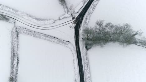 Topdown-of-a-snow-covered-countryside-with-a-cleared-tarmac-road-and-a-junction-splitting-off-covered-in-snow-with-visible-car-tyre-tracks