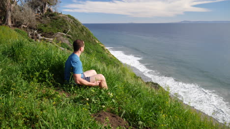Ein-Mann,-Der-In-Beruhigender-Ruhe-Am-Rande-Einer-Strandklippe-Sitzt-Und-Tief-Nachdenkt-Und-Meditiert,-An-Einem-Strahlend-Sonnigen-Tag-In-Santa-Barbara,-Kalifornien,-Zeitlupe