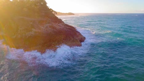 Aerial-view-of-a-rocky-cliff-and-turquoise-blue-water-waves-splashing-at-sunset