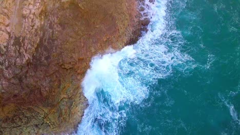 Aerial-shot-of-a-wave-hitting-a-cliff-with-turquoise-blue-water-and-a-lot-of-splashing-seafoam-at-sunset