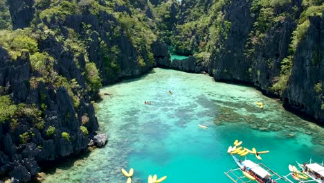 Low-flying-aerial-of-boats-and-kayaks-at-entrance-to-big-lagoon,-small-lagoon,-El-nido,-Palawan,-Philippines