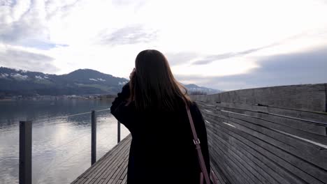 Following-shot-of-a-young-asian-woman-doing-her-hair-while-walking-on-a-wooden-pier-in-slow-motion-in-Rapperswil-Switzerland