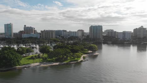 Beautiful-morning-aerial-of-downtown-Sarasota,-lush-green-coastal-park-and-harbor-which-is-home-to-an-enviable-number-of-large-motorboats-and-yachts