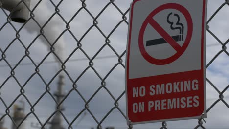 A-refinery-pipe-smoking-and-polluting-in-the-background-pulling-focus-to-a-no-smoking-sign-in-the-foreground-on-a-chain-linked-fence