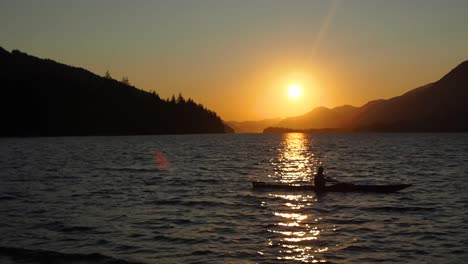 Sonnenuntergang-An-Einem-See-Mit-Bergen-Im-Hintergrund-Und-Einem-Paddle-Boarder,-Der-Durch-Das-Licht-Geht