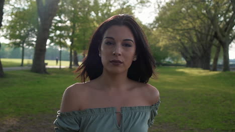 Slow-Motion-Portrait-of-a-gorgeous-hispanic-latino-young-woman-looking-at-the-camera-and-show-emotions-from-serious-to-smiling-and-laughing-with-a-beautiful-British-park-in-the-background
