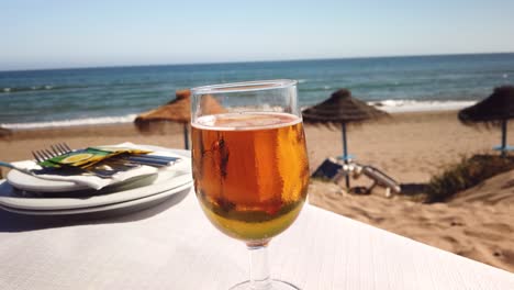 cold-refreshing-beer-glass-on-a-table-with-sea-view-and-a-Spanish-beach-in-Marbella,-filmed-in-4k