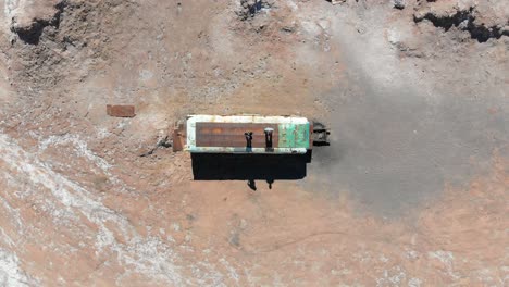 Aerial-top-view-of-an-abandoned-bus-with-two-travelers-on-the-ceiling-in-Atacama-desert,-South-America,-Chile