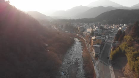 Drone-flight,-over-small-Japanese-town-of-Nikko,-misty-background-during-golden-hour-with-rays-shining-showing-silhouettes-of-mountains-in-the-background-and-a-river-running-through-the-middle