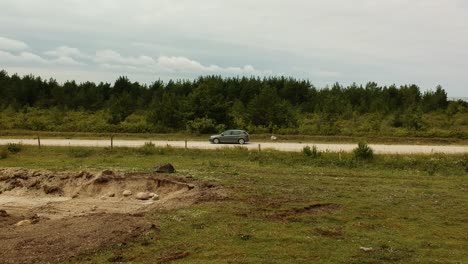 Aerial-side-shot-following-a-grey-car-in-a-open-dusty-straight-road-near-some-trees-in-a-cloudy-day