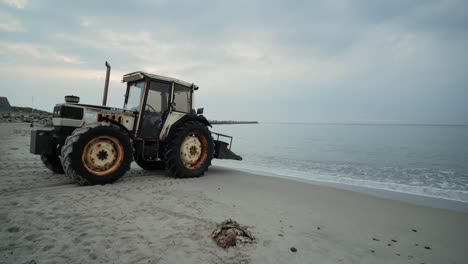 Rusty-tractor-standing-on-sand-at-a-beach-with-waves-and-clouds-and-a-mole-in-the-background